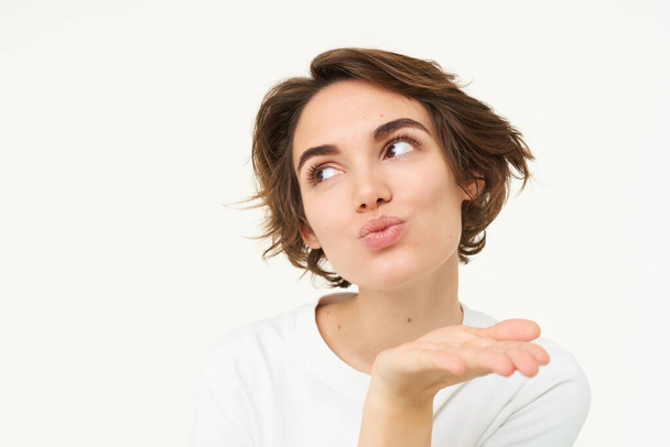 Close up of brunette woman blowing air kiss, mwah, kissing gesture, pucker lips and gazing away, standing over white background. Copy space - Photo, Image