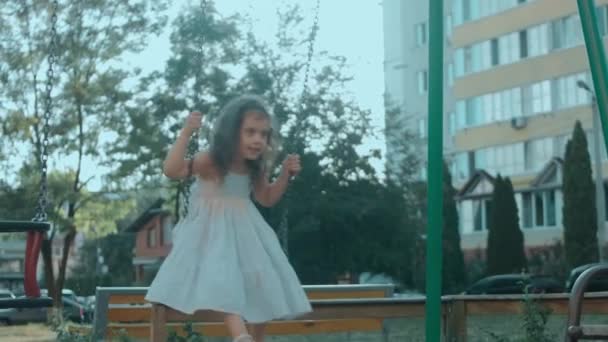 Retrato de uma menina pré-escolar encaracolada feliz jogando em um balanço no playground em um dia ensolarado de verão - Filmagem, Vídeo