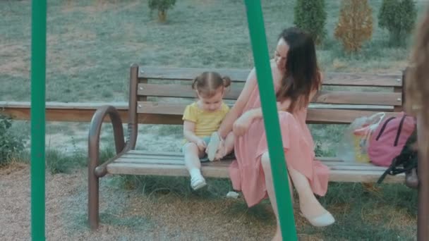 Мама и дочь сидят на скамейке на детской площадке. Кавказская молодая счастливая женщина чистит своим маленьким девочкам обувь из маленьких камней. - Кадры, видео