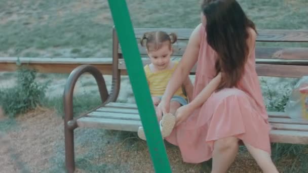 Мама и дочь сидят на скамейке на детской площадке. Кавказская молодая счастливая женщина чистит своим маленьким девочкам обувь из маленьких камней. - Кадры, видео