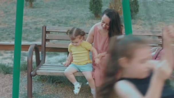 Anne ve kızı oyun parkında bir bankta oturuyorlar. Beyaz, genç, mutlu bir kadın küçük kızını kucağına aldı ama çocuk oynamak istiyor. - Video, Çekim