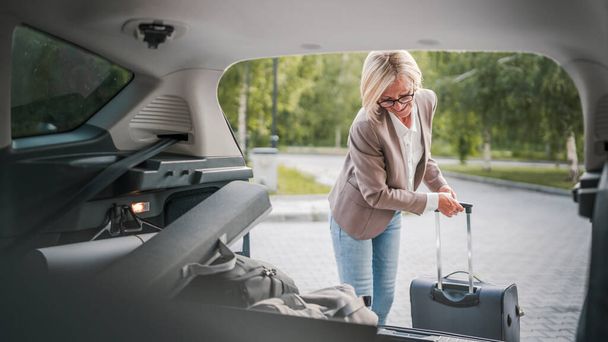 One mature blonde beautiful business woman travel concept female take stuff belongings from the back of her car while moving or arrive to destination or work real people copy space - Photo, Image