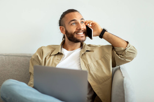 Young stylish guy entrepreneur sitting on couch, working on laptop at home, talking on phone, looking at copy space for advertisement and smiling. Freelance, remote work, home business - Photo, Image