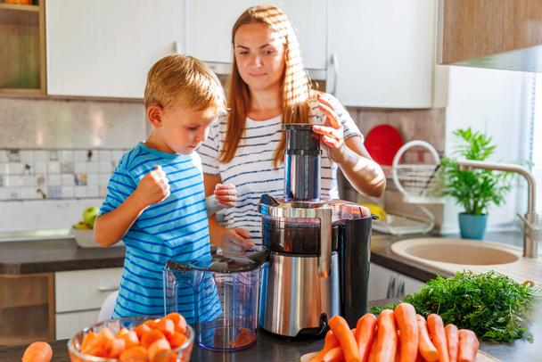 Μικρό παιδί με μητέρα φτιάχνει φρέσκο χυμό καρότου με αποχυμωτή σε μια ηλιόλουστη κουζίνα - Φωτογραφία, εικόνα