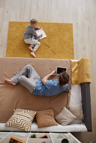 High angle view of young father lying on bed and surfing net on tablet while son sitting on carpet and drawing on paper - Photo, image