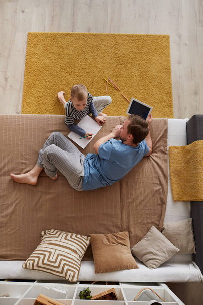 Above view of little blond-haired boy sitting at sofa bed and making picture with father who surfing internet at tablet - Photo, image
