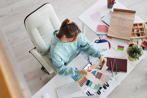 Above view of creative young designer sitting at table with massy sketches and choosing colors for new dresses - Фото, изображение