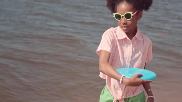 Medium slow mo van mooie kleine zwarte meisje in een zonnebril staande enkel diep in het water gooien frisbee - Video