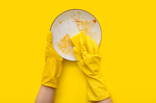 Ženské ruce v gumových rukavicích mytí špinavý talíř s houbou na žlutém pozadí - Fotografie, Obrázek
