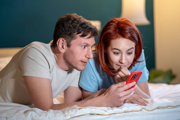 Ευτυχισμένο ζευγάρι Lyimg γάμου στο κρεβάτι μιλώντας ανάγνωση καλά νέα στην οθόνη του τηλεφώνου. Ενθουσιασμένη σύζυγος και σύζυγος λαμβάνουν σημαντικό μήνυμα στο κινητό μαζί. Περήφανος άνδρας και γυναίκα έκπληκτος κερδίσει σε απευθείας σύνδεση λαχείο - Φωτογραφία, εικόνα