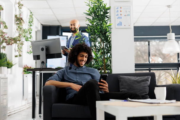 Uśmiechnięty arabski lider zespołu startującego omawia projekt ze współpracownikami na internetowym spotkaniu na smartfonie. Pracownik biznesu na czacie w telefonie komórkowym videocall podczas siedzenia w przestrzeni coworking - Zdjęcie, obraz