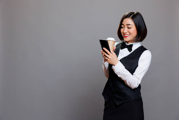 Sorrindo mulher asiática vestindo uniforme recepcionista mensagens de texto e rolagem de mídia social enquanto bebe café para ir. Restaurante garçonete alegre desfrutando de chá em caneca de papel e usando telefone celular - Foto, Imagem