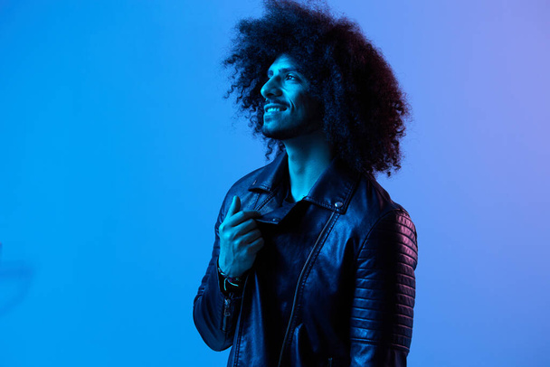 Portrait of fashion man with curly hair on blue background multinational, colored light, black leather jacket trend, modern concept. High quality photo - Photo, Image