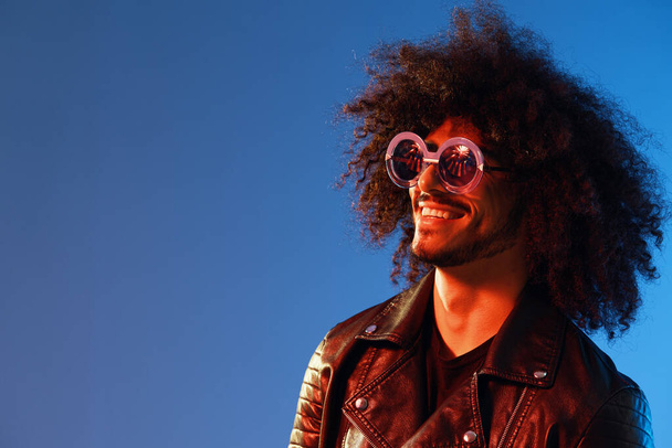 Portrait of a stylish man with curly hair with glasses smile on blue background multinational, color light, black leather jacket trend, modern concept. High quality photo - Foto, Bild