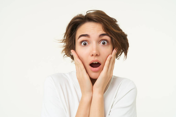 Portrait of girl with surprised face, makes shocked expression, stands over white background. Emotions and people concept - Photo, Image