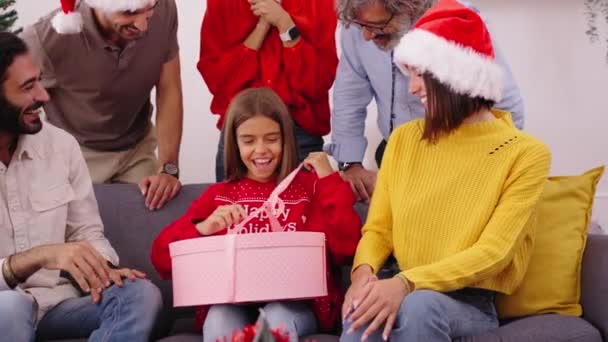 Emocionado niño alegre abriendo regalo de Navidad sentado en casa. Riendo sorprendió a la familia que llevaba sombreros de Santa Claus reunidos para el intercambio de regalos. Relaciones domésticas positivas en vacaciones - Imágenes, Vídeo