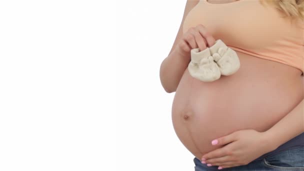 Kobieta w ciąży umieścić Baby botki na brzuchu. Zbliżenie białych butach dziecięcych na brzuch w ciąży. Ciężarne kaukaski głaszcząc jej brzuch na białym tle - Materiał filmowy, wideo