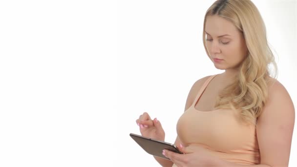 タブレットを持っている妊娠中の白人女性のスタジオショット。妊娠中の女性は、白い裏地にタブレットを使用しています。タブレットのタッチスクリーン上の妊娠中のブロンドのタイピング - 映像、動画