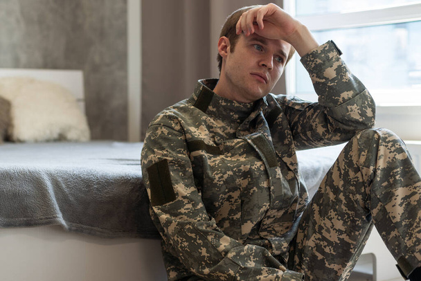 Gestresste veteraan soldaat verdiept in depressieve gedachten en herinneringen. Droevige depressieve jonge militaire man in camouflageuniform zittend op de bank en denkend aan oorlog. PTSS en therapie concept - Foto, afbeelding
