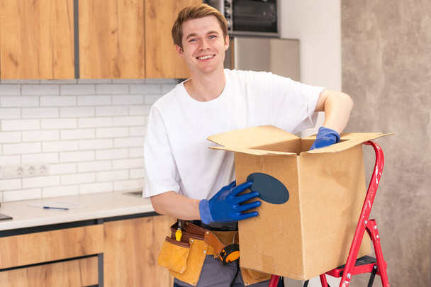 Smiling male worker of moving and delivery company holding cardboard box. Loader in overalls posing against background of colleague who packs cardboard boxes. Moving service concept - Photo, Image