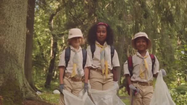 Střední portrét multietnického skautského oddílu žáků základního věku stojících s pytli na odpadky venku ve sluncem zalitém zeleném letním lese, pomáhajícím prostředí - Záběry, video