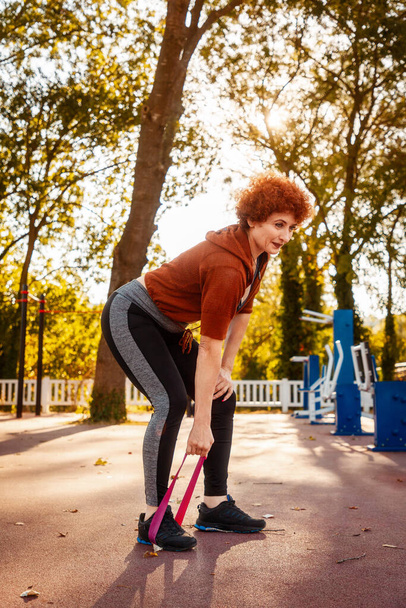 Mujer caucásica adulta hace estiramiento con banda elástica en la calle. Entrenamiento en el parque. Orientación vertical. Concepto de estilo de vida saludable. - Foto, imagen