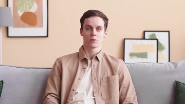 Vyötärö ylös POV nuori valkoihoinen mies puhuu kameran edessä istuu sohvalla olohuoneessa pastelli persikka seinät - Materiaali, video