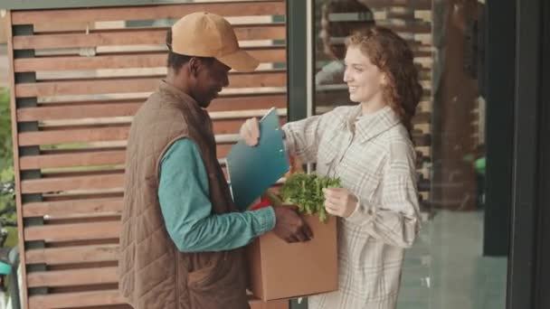 Середній знімок усміхненої молодої кавказької жінки, що зустрічається з чорношкірим чоловіком, який доставляє продукти харчування на її місце - Кадри, відео
