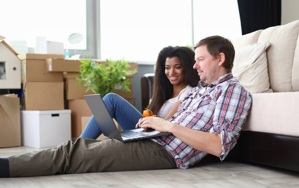 Retrato de casal sentado no tapete macio no novo apartamento. Mulher e homem entretidos com laptop moderno. Caixas vazias no quarto. Dia comovente e conceito imobiliário
 - Foto, Imagem