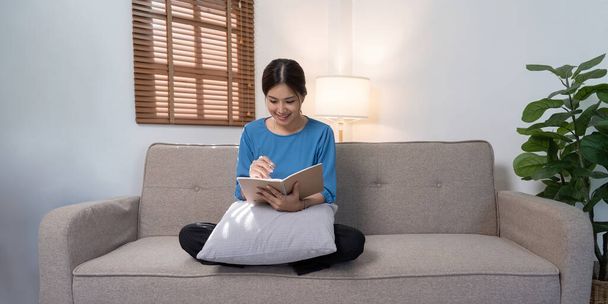 Hermosa joven asiática sentada y tomando nota en el sofá de la casa. Feliz sonriente trabajo en línea, trabajo independiente en casa. - Foto, imagen