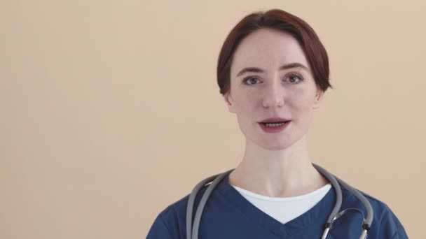 Chest up POV mladé bělošky ošetřovatelky v křoví a modrý stetoskop na ramenou dívá a mluví na kameru stojící proti pastelové broskvové stěně uvnitř - Záběry, video