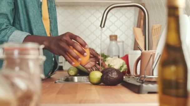 Καλλιεργημένη φωτογραφία αγνώριστου μαύρου άνδρα να πλένει φρέσκα λαχανικά στο νεροχύτη στην κουζίνα ενώ μαγειρεύει - Πλάνα, βίντεο