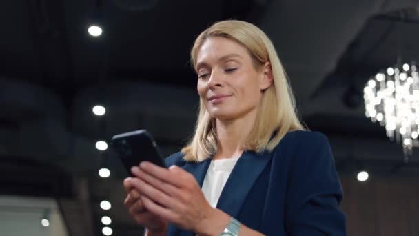 Low angle view of pleasant lady typing message on modern smartphone while standing at hall of office building. Smiling businesswoman using modern gadget for communicating with coworkers online. - Footage, Video