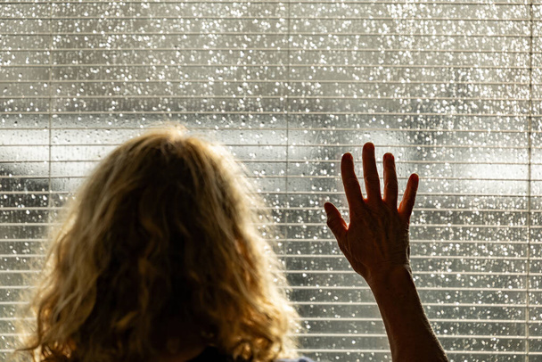 Kobieta spogląda przez jasne okno z żaluzjami podczas letniej burzy deszczowej ze słońcem. - Zdjęcie, obraz