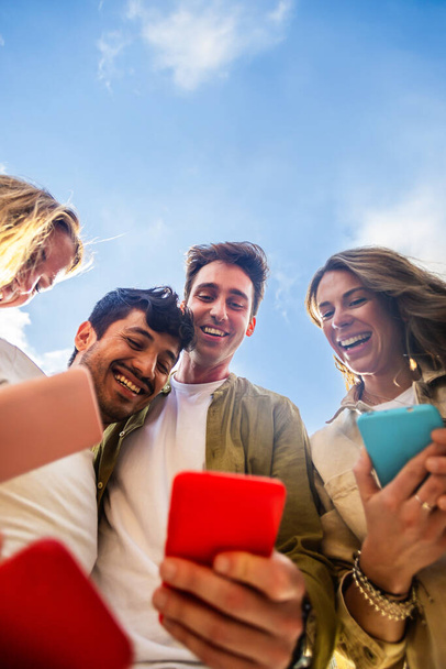 Pystysuora laukaus ryhmä ihmisiä käyttävät matkapuhelin laite seisoo yhdessä ympyrässä ulkona. Millennial ystävät riippuvainen sosiaalisen median sovellus, vedonlyönti tai videopelin pelaaminen alustalla verkossa. - Valokuva, kuva