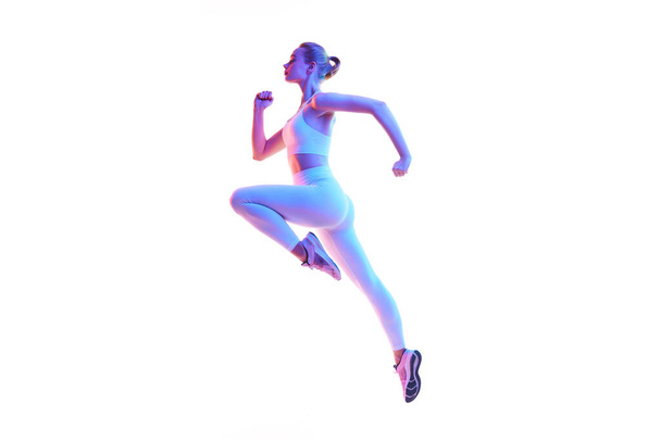 Retrato de visão lateral da jovem mulher esportiva correndo em ação isolada contra fundo branco em luz de néon. A aquecer. Conceito de esporte, fitness, estilo de vida saudável, força, juventude, anúncio. Espaço de cópia - Foto, Imagem
