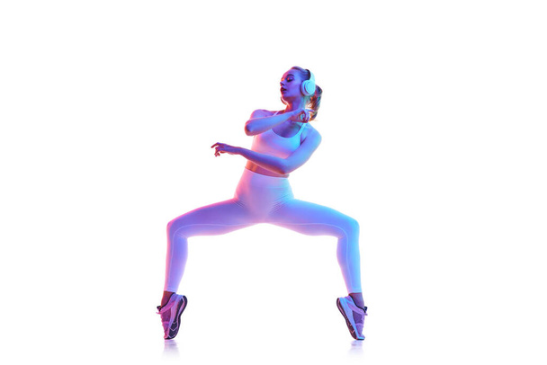 Retrato de vista lateral da jovem mulher magra em plie pose com música em fones de ouvido fundo branco isolado em luz de néon. Conceito de esporte, fitness, estilo de vida saudável, força, juventude, anúncio. Espaço de cópia - Foto, Imagem