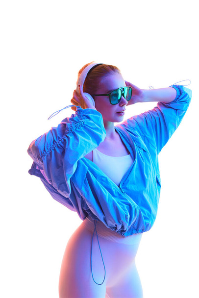 Retrato de chica delgada vestida con ropa deportiva de moda con gafas de sol y auriculares en luz de neón sobre fondo blanco. Concepto de estilo de vida activo, deporte, moda, movimiento. Copiar espacio - Foto, imagen