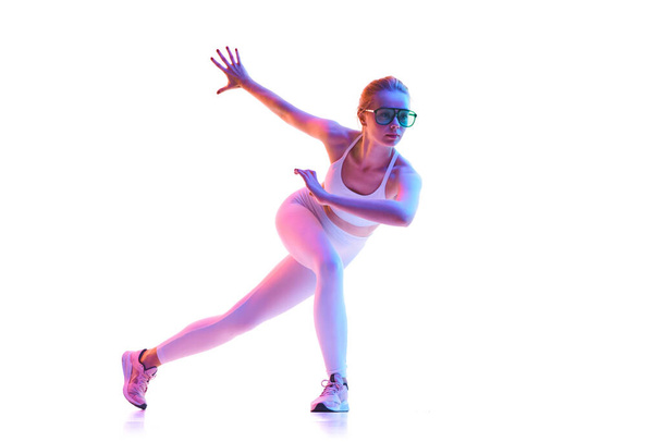 Takana näkymä ohut tyttö pukeutunut muoti asu aurinkolasit tehdä harjoituksia jalat neon valoa valkoisella taustalla. Käsite aktiivinen elämäntapa, urheilu, terveys, kilpailu. Kopioi tila - Valokuva, kuva