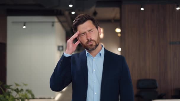 Tired handsome caucasian man massaging temples with hands and trying to relieve pain in head while standing in office. Bearded male feeling bad after hard working day in modern business center. - Footage, Video