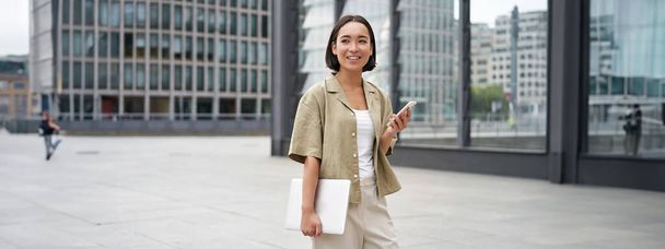 Dizüstü bilgisayarı ve akıllı telefonu olan Asyalı bir kız şehir merkezinde kameraya gülümsüyor.. - Fotoğraf, Görsel
