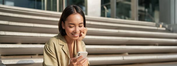アジアの女性の垂直ショット,学生は、市内の階段に座っています,携帯電話の画面を見て、笑顔,スマートフォンアプリを使用して. - 写真・画像