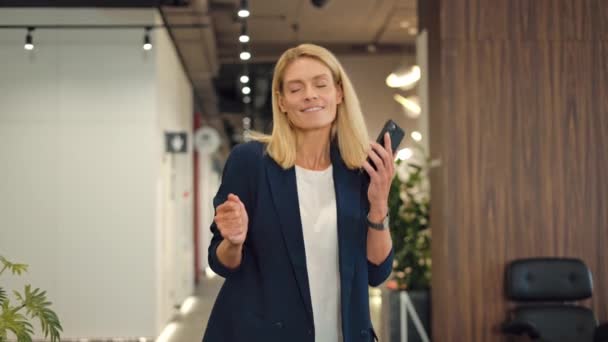 Female financial director taking break from work and dancing at coworking space with mobile phone in hand. Cheerful fair-haired lady in elegant attire relaxing to cool music in empty corridor. - Footage, Video