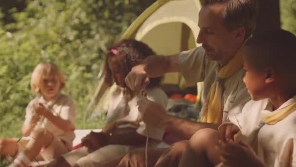 Skilled mid aged Caucasian male scouter teacher diverse cub scouts to make ties while camping together in summer forest - Footage, Video