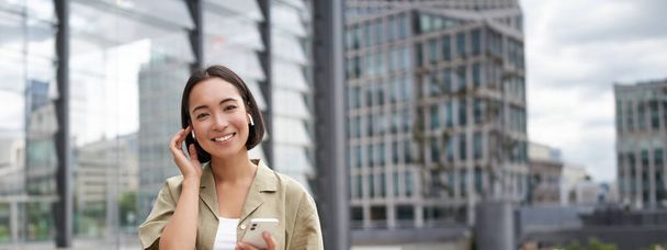 Joven mujer asiática sonriente en auriculares inalámbricos, camina por la calle con teléfono inteligente y auriculares. - Foto, Imagen