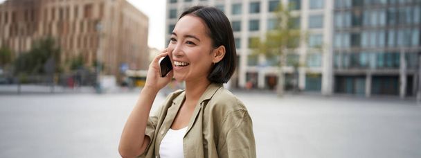 Modern Asyalı genç kız cep telefonuyla konuşuyor, şehir caddesinde telefon kullanıyor. Akıllı telefondan birini ararken gülümseyen bir kadın.. - Fotoğraf, Görsel