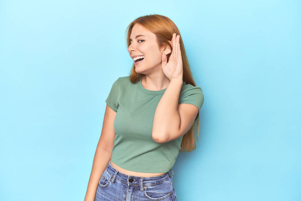 Κοκκινομάλλα νεαρή γυναίκα σε μπλε φόντο προσπαθεί να ακούσει ένα κουτσομπολιό. - Φωτογραφία, εικόνα