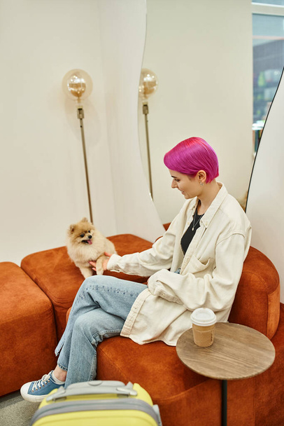 κομψή γυναίκα παίζει με το δικό του σκυλί, ενώ κάθεται σε ξενοδοχείο συντροφιάς κοντά σε ταξιδιωτική τσάντα και καφέ για να πάει - Φωτογραφία, εικόνα
