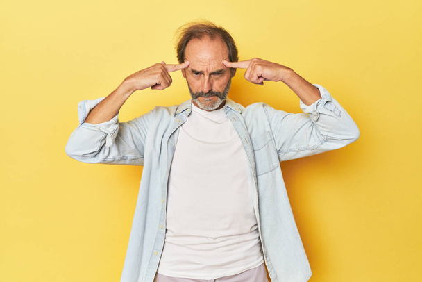 Blanke man van middelbare leeftijd in gele studio gericht op een taak, het houden van wijsvingers wijzend hoofd. - Foto, afbeelding