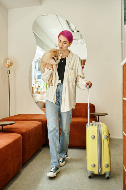 mujer de moda con pelo morado sosteniendo spitz pomeraniano y caminando con maleta en hotel de mascotas - Foto, Imagen